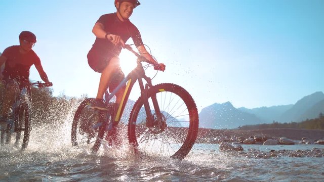 SUPER SLOW MOTION LENS FLARE LOW ANGLE: Water drops fly around two guys riding electric bicycles along Soca river on a beautiful spring day. Blinding sunbeams shine on mountain bikers splashing water