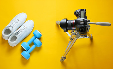 Minimalistic sport concept. Fitness blogging. White sneakers with plastic dumbbells and camera with tripod on yellow background. Top view