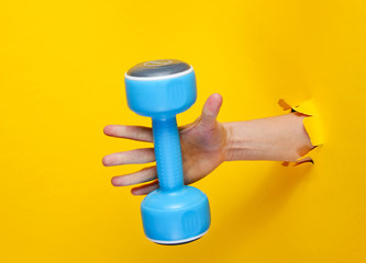 Female hand takes a blue plastic dumbbell through torn yellow paper. Minimalistic sport concept
