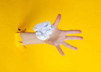 Fototapeta na wymiar Female hand throws white crumpled ball of paper through the torn yellow paper background. Minimalistic idea concept