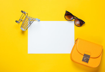 White sheet of paper for copy space, mini shopping trolley, bag, sunglasses on yellow background. Creative shopping background - Powered by Adobe