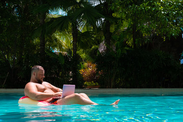 male with an inflatable ring in the water in the pool works on the computer. Funny Fat Man in Swimming Circle with Pink Laptop. freelance concept