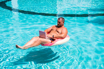 Freelancer with an inflatable ring in the water in the pool works on the computer. Funny Fat Man in Swimming Circle with Pink Laptop. freelance concept