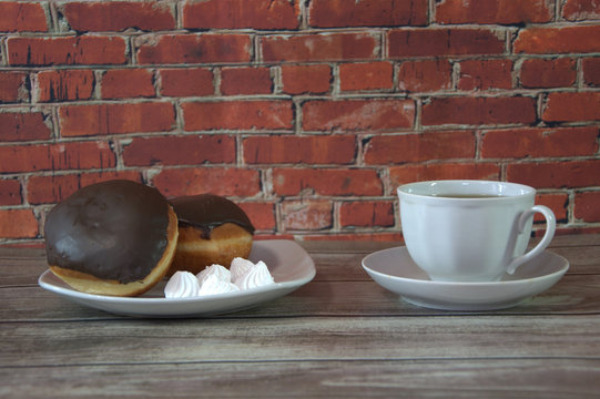 A cup of black tea on a saucer and a plate with two donuts in chocolate icing and pieces of bize, lie on a wooden table against a brick wall. Close-up.