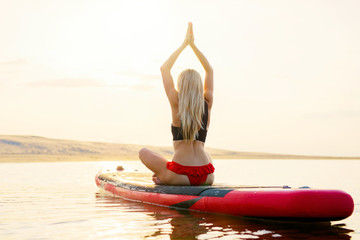 Fit woman doing yoga exercises on paddle board in the water at sunset