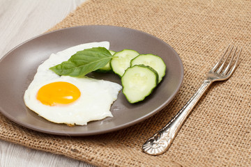 Plate with fried egg, fresh cut cucumber and leaf of basil on sackcloth.