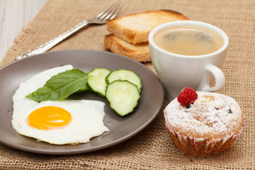 Fototapeta premium Plate with fried egg, fresh cut cucumber and leaf of basil, fork, cup of black coffee, cupcake and toasts.