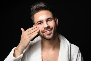 Handsome man applying clay mask on his face against dark background