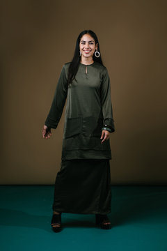 Studio portrait of an elegant, beautiful, tall, slim and attractive Malay Asian woman in a traditional green silk baju kurung dress against a brown background. She is dressed for visiting during Raya.