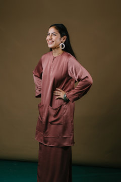 Portrait of a tall, slim and elegant Muslim woman in a salmon pink silk traditional dress (baju kurung) in a studio. She is smiling and looks confident as she has her hands on her hips.