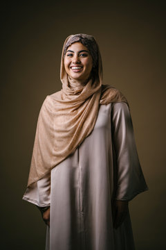 A young and beautiful Muslim Middle Eastern woman in a brown traditional outfit and hijab head scarf smiles as she poses in a studio against a tan background. She is wearing a pearl earring.