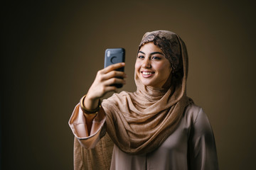 Fototapeta premium A young and beautiful Muslim woman in a traditional outfit and hijab head scarf is looking at her smartphone to take a selfie of herself in a studio. She is smiling happily as she poses for her photo