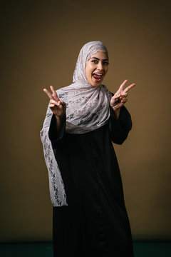 Portrait of a tall, slim and elegant Muslim Middle Eastern woman in a black dress and white hijab head scarf smiling and laughing as she makes a peace sign with her fingers. She is having fun.