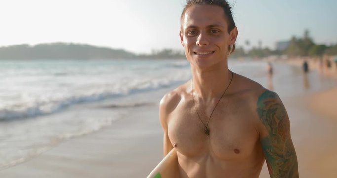 Portrait of a surfer full of strength and confidence. A guy stands with a board on the beach surrounded by palm trees.