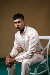 Fototapeta premium Portrait of a young, handsome and athletic Malaysian Indian Muslim man in a cream colored traditional baju melayu tunic sitting on a chair. He is dressed festively to go visiting for Raya. 