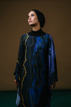 Studio portrait of a tall, slim, elegant and beautiful Middle Eastern Muslim woman in a flowing traditional blue dress with a turban head scarf. She is dressed to celebrate Raya. 