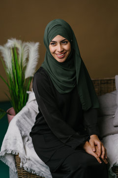 Portrait of a beautiful, elegant and attractive young Middle Eastern Muslim woman in a black dress and an olive green hijab head scarf. She is smiling as she sits on a couch at home and relaxing. 