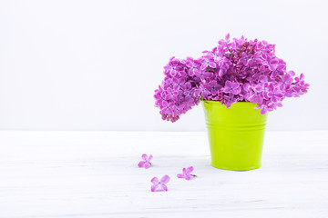 Blooming lilac bouquet in green bucket shaped vase on white wooden background.