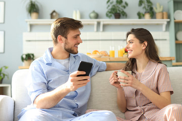 Young couple watching online content in a smart phone sitting on a sofa at home in the living room.
