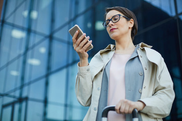 Below view of serious businesswoman in eyeglasses standing against modern glassy building and using gadget while reading message on move