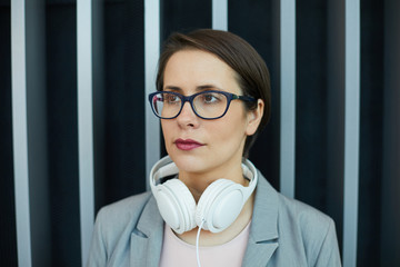 Close-up of serious pensive young businesswoman with headphones on neck wearing eyeglasses...