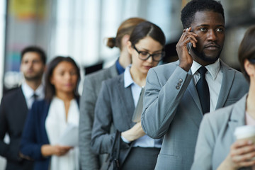 Serious displeased young African-American businessman with beard standing in line and calling on...