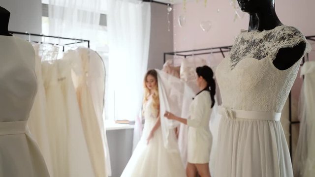 Wedding consultant helps bride choosen wedding dress in salon and wearing it near large mirror