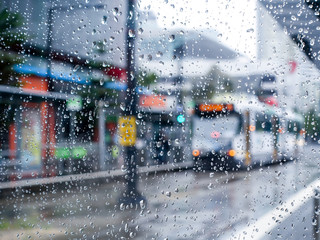 Close up of raindrops on wet glass window of a tram stop in city. Blurry view of a tram stopping at platform with shelter to take passengers on a rainy day. Melbourne, VIC Australia.