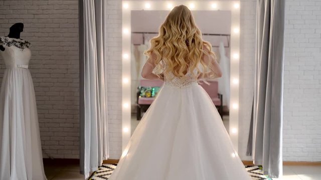 beautiful blonde curly girl  in wedding salon is wearing a dress near loft mirror