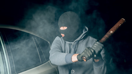 robber in a Balaclava tries to break the car glass with a baseball bat