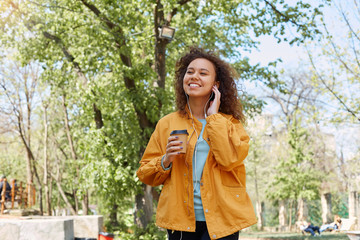 Young attractive dark skinned curly girl broadly smiling, wearing a yellow jacket, drinking coffee, holding headphones with hand and enjoying favorite music walking in the park in fine weather.