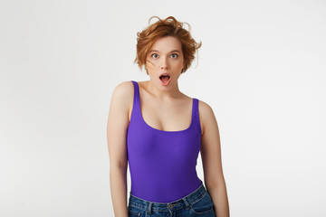 Young surprised attractive short-haired girl, wearing a purple jersey, looking at the camera in surprise with wide open mouth and eyes, she heard the shocking news. Isolated over white wall.