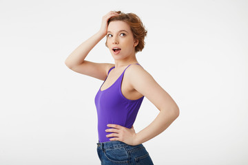 Portrait of hocked attractive short-haired girl, wearing a purple jersey, looking away in surprise with wide open mouth and eyes, straightens hair with his hand, isolated over white wall.