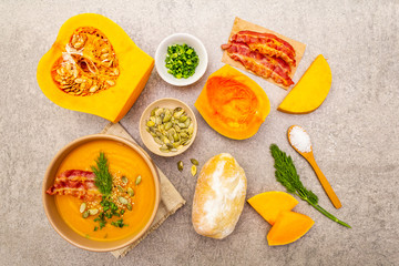 Homemade autumn hot pumpkin cream soup with smocked bacon and seeds. Raw pumpkin, bun, fresh chives, dill, salt in spoon, vintage linen cloth on stone background, top view