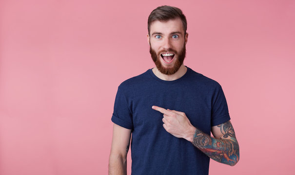 Young happy amazed attractive red-bearded young guy , wearing a blue t-shirt, with wide open mouth in surprise, pointing finger to copy space at the left side isolated over pink background.