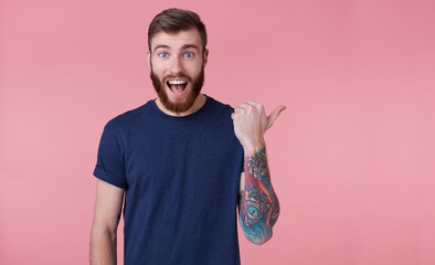 Young happy red-bearded young guy , with wide open mouth in surprise, saw something wrong, wants to draw your attention pointing finger to copy space at the right side isolated over pink background.