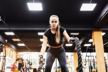 Portrait of a beautiful positive hardy young woman fitness model engaged with ropes in the gym. The...
