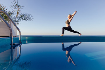 Young attractive woman practicing yoga stand in Warrior one exercise, Virabhadrasana pose, working out by the pool, above the beach, relaxing against blue sky. Health and beauty concept. Copy space