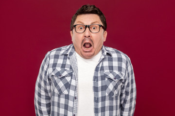 Portrait of shocked handsome middle aged business man in casual checkered shirt and eyeglasses standing and looking at camera with open mouth and big eyes. studio shot, isolated on dark red background
