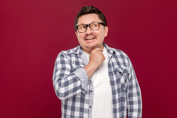 Fototapeta premium Portrait of smiley pondering handsome middle aged business man in casual checkered shirt and eyeglasses standing looking away and dreaming. indoor studio shot, isolated on dark red background.