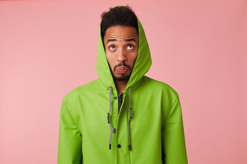 Close up of wondered young African American handsome guy in green rain coat, looks up in confused, clenched lips, stands over pink background.