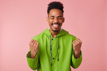 Joyful young african american handsome guy stands over pink background, clenched his fists, broadly smiling and absolutely happy - he won the lottery!