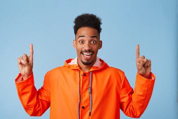 Hey, look up! Happy young attractive african american man wears in orange rain coat, smiles broadly stands over blue background and wants to draw your attention at copy space, points fingers up.