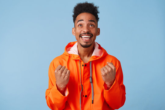 Young Attractive African American Dark Skinned Guy Wears In Orange Rain Coat, Feels Very Happy, Smiles Broadly, Look Up And Clenches His Fists Stands Over Blue Background.