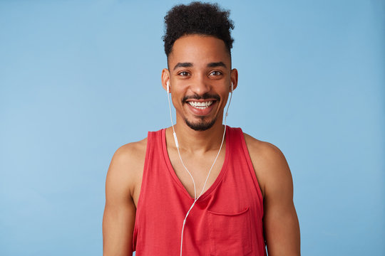 Glad young african american handsome man feels great, wears in red shirt, listens to a new exciting podcast, broadly smiles and looks in camera stands over blue background.