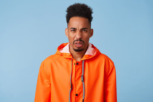 Close up of uhappy young african american dark skinned man in disgust, dissatisfied with the rainy weather outside, wears in orange rain coat, frowns isolated over blue background.
