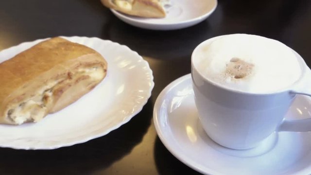White mug with hot cappuccino coffee on the table. raisin bun lying on a plate. Mug in focus