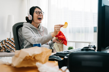 happy asian female nerd holding bag of chip snack junk food with trash on desk looking monitor...