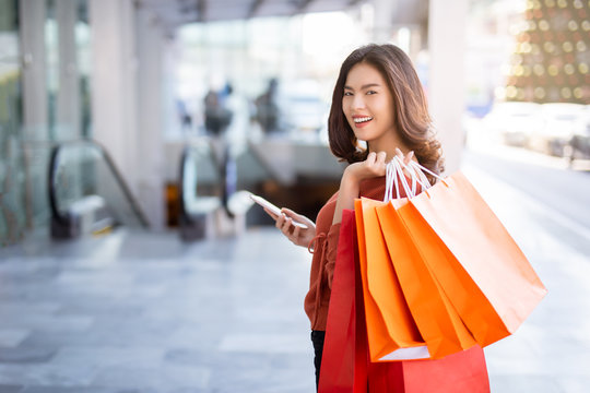 Happy Asian pretty girl holding shopping bags while looking at smart phone at shopping mall concept.