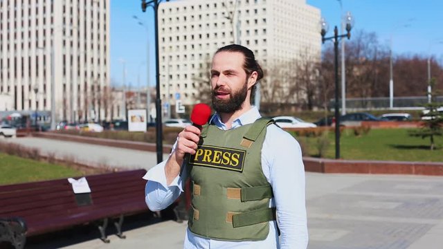 Medium shot, male television journalist in a bulletproof jacket talking to a microphone in front of skyscrapers in the financial district of the city.